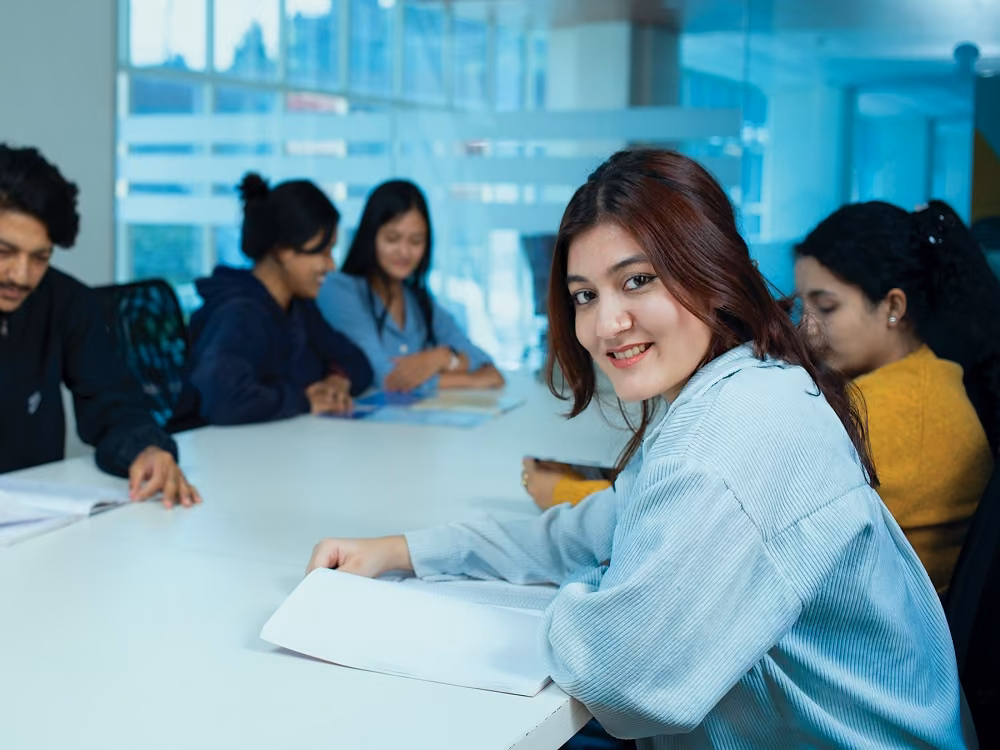 Students round a meeting table with papers, a glass-walled room behind them