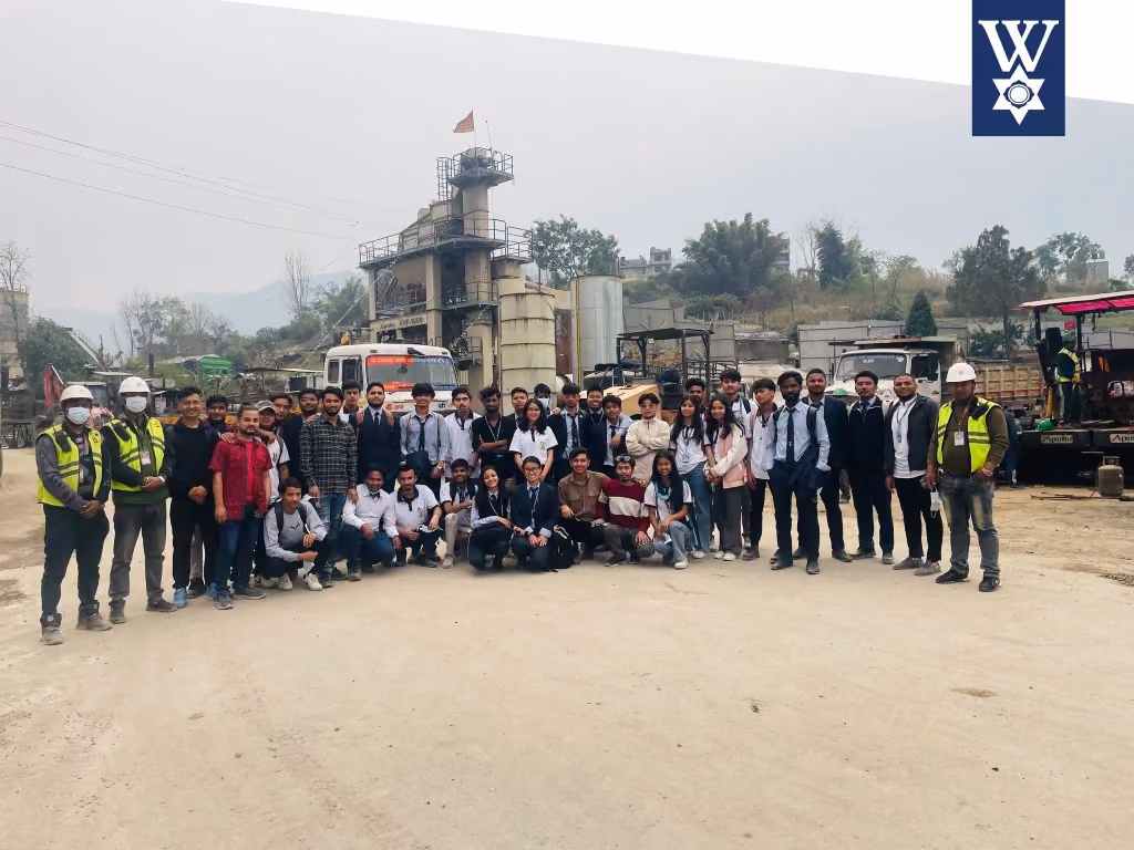 A large student group in hi-vis at an asphalt plant, machinery and hills behind them