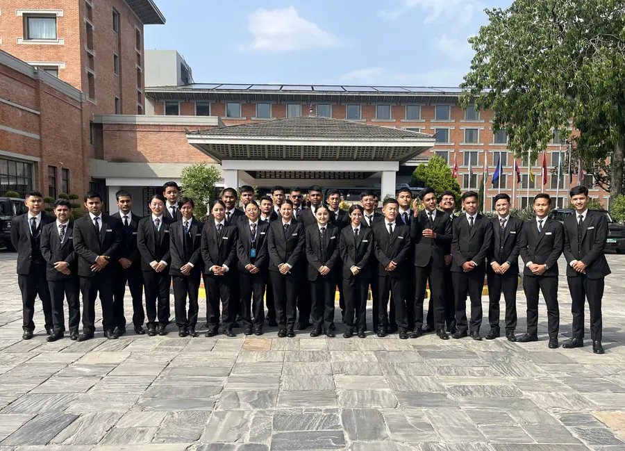 A large group of students in blazers outside the brick block, the flagpoles behind them