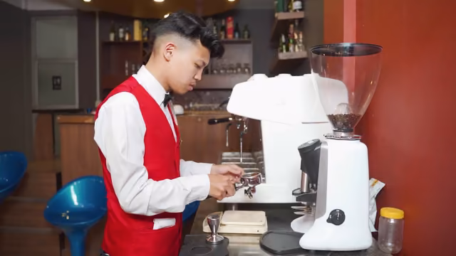 A student in waistcoat and bow tie working the espresso machine and grinder at the training bar