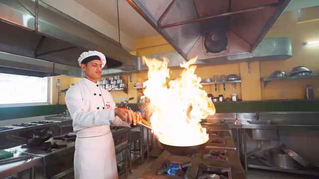 A student in chef's whites tossing a flaming wok under the extractor hood of the training kitchen